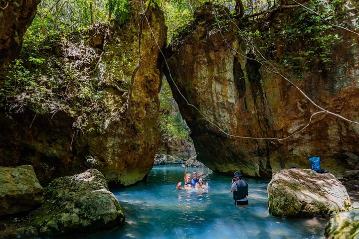People swimming and taking photos in the blue water among large canyon boulders.