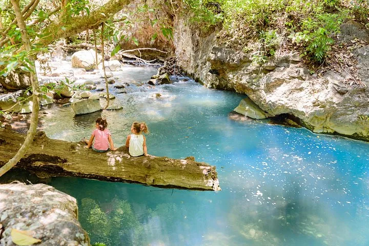 Two young girls sitting on a log over a bright blue river.