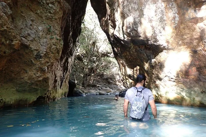 Mann mit wasserdichter Tasche, der in einem felsigen Canyon durch blaues Wasser watet.
